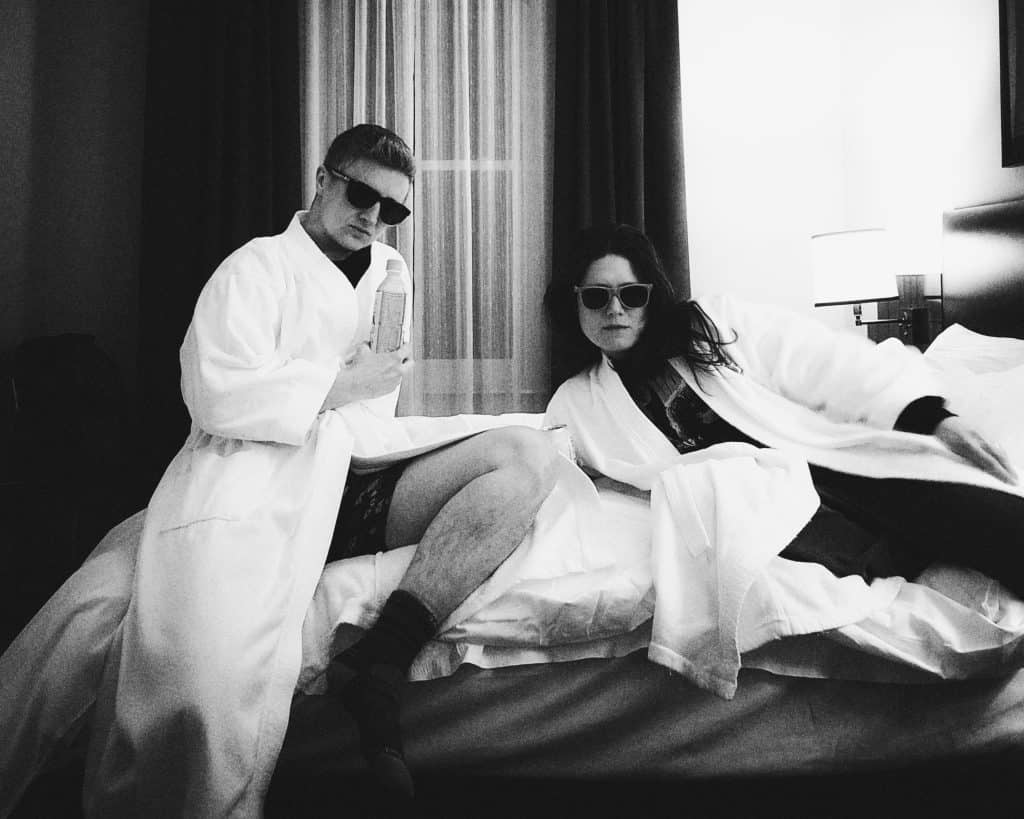Emily and Meg recline on a hotel bed, both dressed in fluffy white bathrobes and wearing sunglasses.